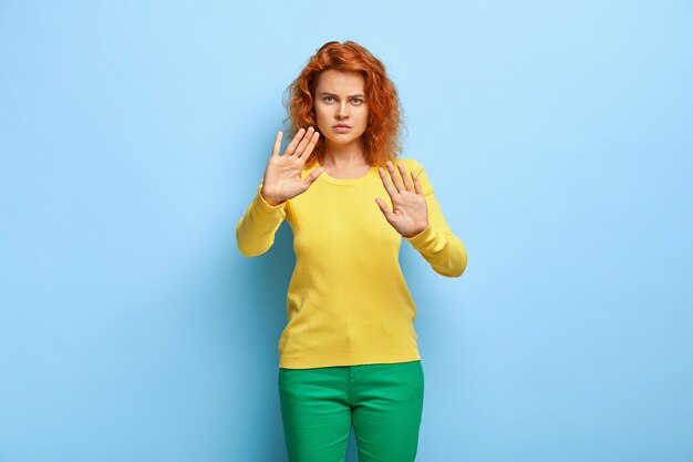 Serious discontent woman has wavy red hair, shows stop gesture, keeps palms outstretched at camera, refuses something