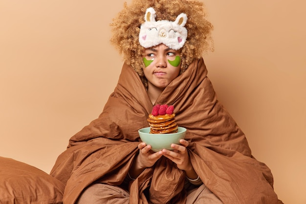 Foto grátis mulher séria descontente com cabelo encaracolado enrolado em cobertor macio segura tigela de panquecas com morango vai tomar café da manhã apetitoso acorda de manhã cedo isolado sobre fundo bege