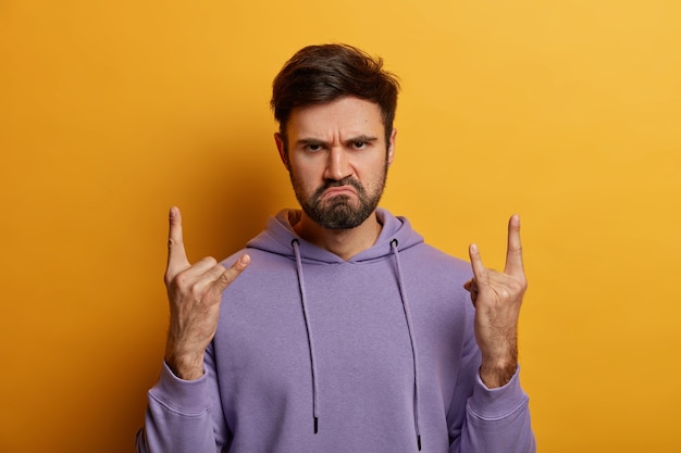 Serious displeased bearded man rocker makes horn sign with fingers, has charismatic face expression, frowns face, wears purple sweatshirt, attends rock concert, isolated over yellow wall.