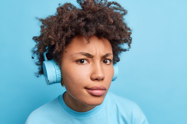 Serious displeased curly haired African American woman wears stereo wireless headphones looks attentively  listens music or audio book dressed casually isolated over blue wall.