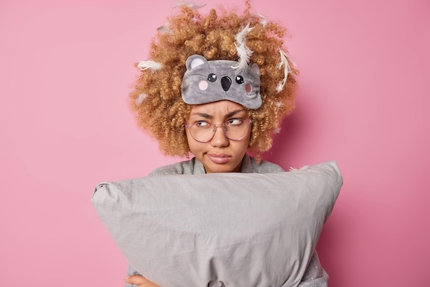 Foto gratuita una donna dai capelli ricci seria e dispiaciuta tiene il cuscino guarda scontento indossa una maschera per dormire sulla fronte ha piume bloccate in pose di capelli ricci su sfondo rosa esprime emozioni negative