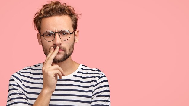 Serious displeased man frowns face, keeps hand on mouth, has curly hair and stubble, wears striped t shirt, round spectacles