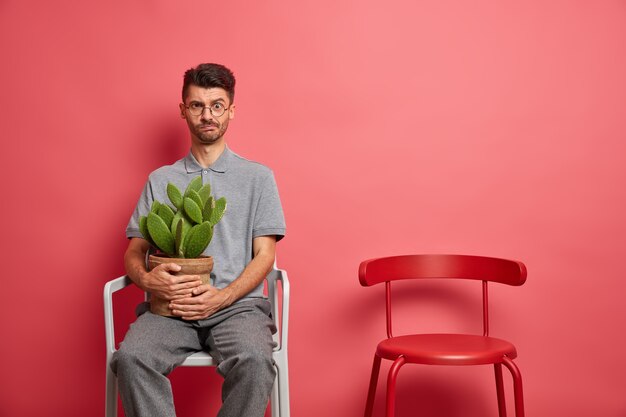 Serious dissatisfied Caucasian man in casual wear rests in chair holds potted cactus stays at home