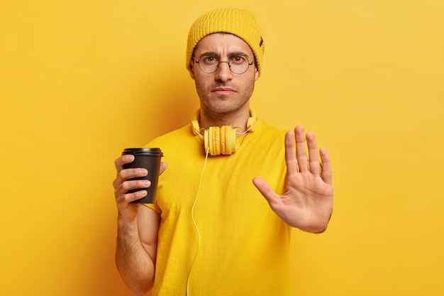 Serious dissatisfied guy makes stop gesture, refuses doing something, says no, holds takeout coffee cup, wears eyewear, yellow casual clothes
