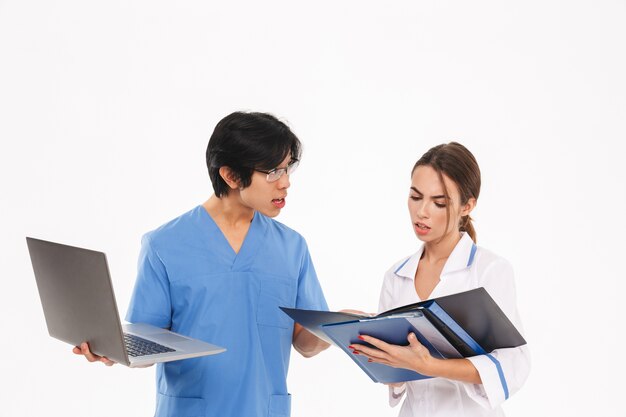 Пара серьезных врачей в униформе, стоя изолированной над белой стеной, работая с портативным компьютером и рентгеновским снимком