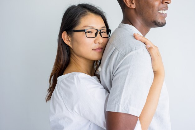 Serious dreamy Asian girl embracing boyfriend from back and looking away. 