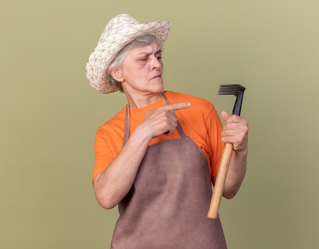 Free photo serious elderly female gardener wearing gardening hat holding and looking at rake