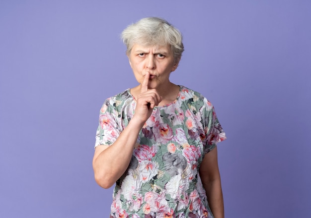 Free photo serious elderly woman puts finger on mouth gesturing hush quiet sign isolated on purple wall