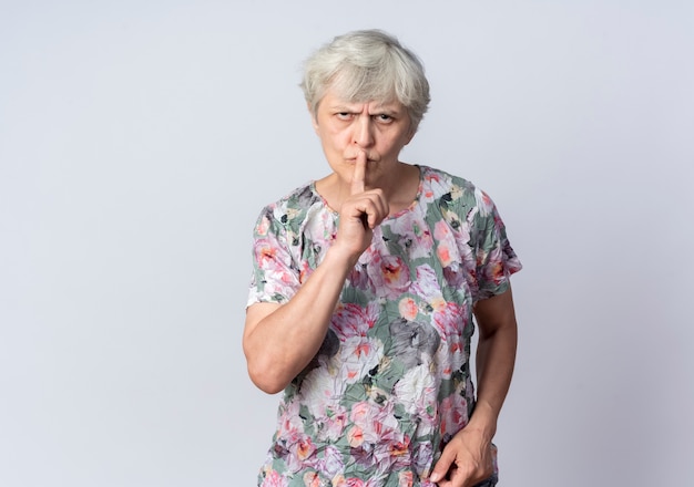 Серьезная пожилая женщина кладет палец на рот, жестикулируя тихий тихий знак на белой стене