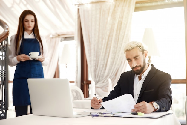 Serious entrepreneur is scanning thefinancial reviews  and the waitress is bringing him hot drink