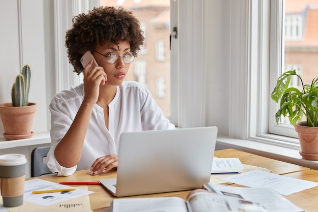 Serious ethnic female CEO wears round spectacles, tries to reach decision with colleague via cell phone