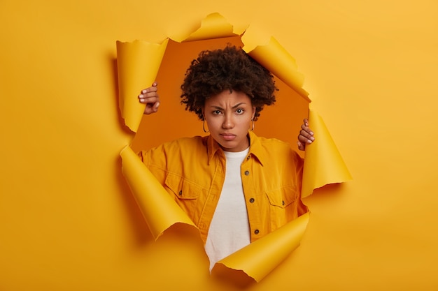 Serious ethnic woman frowns from displeasure, raises eyebrows, dissatisfied with something, wears fashionable denim jacket, stands in paper hole, being distressed and frustrated.