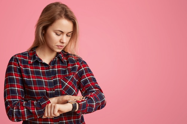 Serious European woman hurries on meeting, looks at watch, checks time.