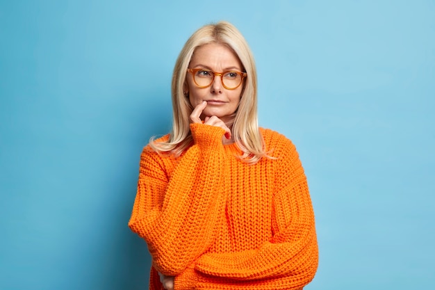 Serious European woman looks thoughtfully considers idea decides on something had doubts dressed in knitted orange jumper.