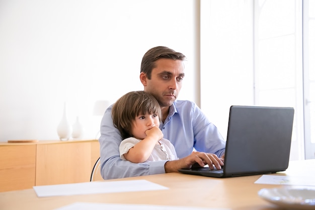 Serious father browsing internet on laptop and holding cute son. Caucasian middle-aged man wearing shirt, sitting at table with kid, looking at screen and working. Fatherhood and home concept