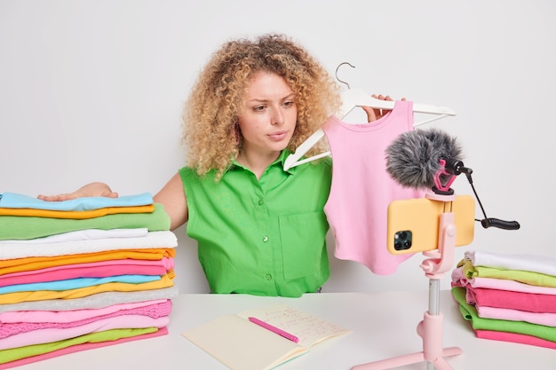 Free photo serious female blogger involved in distance purchasing shares new brand clothes holds pink shirt on hanger records live stream video poses around neatly folded laundry sells outfit through e store