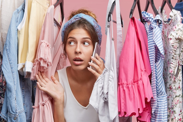 Serious female chatting over mobile phone while looking through rack with clothes, taking advice in her best friend what to wear on date with her boyfriend, having special occasion. Clothing concept