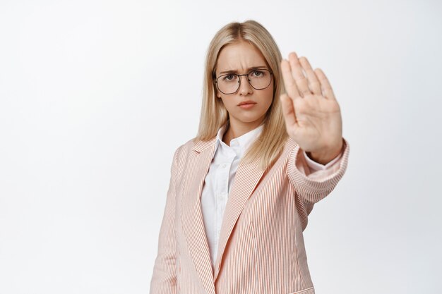 Serious female entrepreneur stretching out hand in stop sign, prohibit, forbid something, saying no, standing on white in suit and glasses