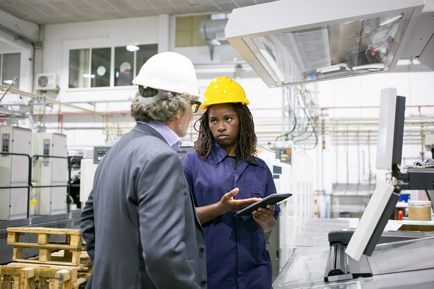 Serious female factory worker talking to boss at machine on plant floor, pointing at tablet screen