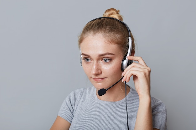 Serious female helpline operator uses headphones for her work, being focused on something, isolated on grey. Businesswoman phones partners. Business, call centre and technology concept