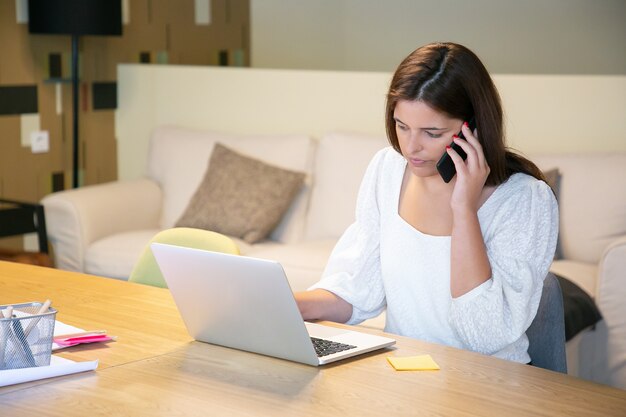 Serious female manager discussing project with customer on phone, sitting at table with laptop and blueprints and typing