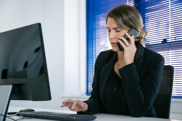 Serious female professional talking on mobile phone while using computer at workplace in office. Medium shot. Digital communication and multitasking concept