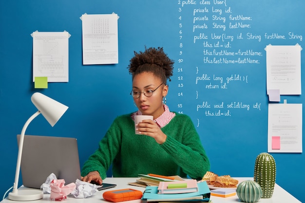 serious female programmer expert works on freelance project in coworking space, drinks coffee, wears round spectacles and green jumper.