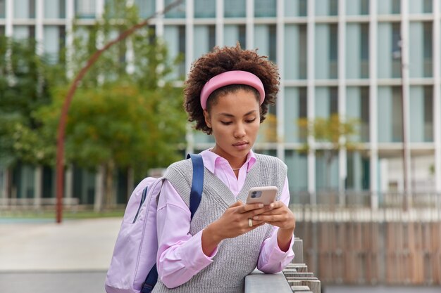 真面目な女子学生が携帯電話でチャットをオンラインで閲覧カジュアルな服装でインターネットを閲覧する現代の都市の建物に対してリュックサックのポーズを外に運ぶ講義後に大学から行く