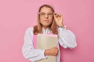 Бесплатное фото Серьезная студентка смотрит через прозрачные очки, одетые в белую блузку, позирует с ноутбуками в помещении, собирается на занятия в университете, изолированные на розовом фоне. изучение концепции