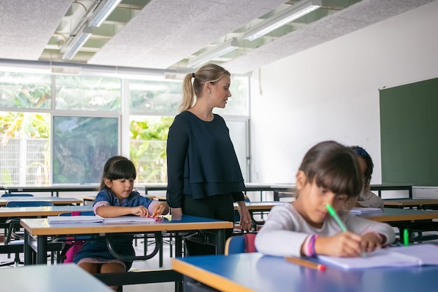 초등학교 아이들이 수업 시간에 책상에 앉아 카피 북에 글을 쓰는 것을 보는 심각한 여성 교사