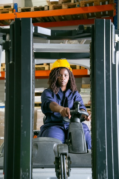 Free photo serious female warehouse worker in hardhat driving fork lift, looking at front