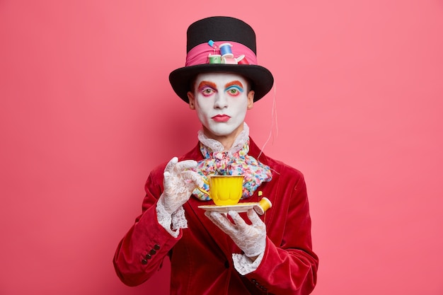Серьезный вымышленный персонаж-шляпник с красочным макияжем пьет кофе с белым лицом, одетым в костюм на хэллоуин, серьезно смотрит в камеру, изолированную на розовой стене