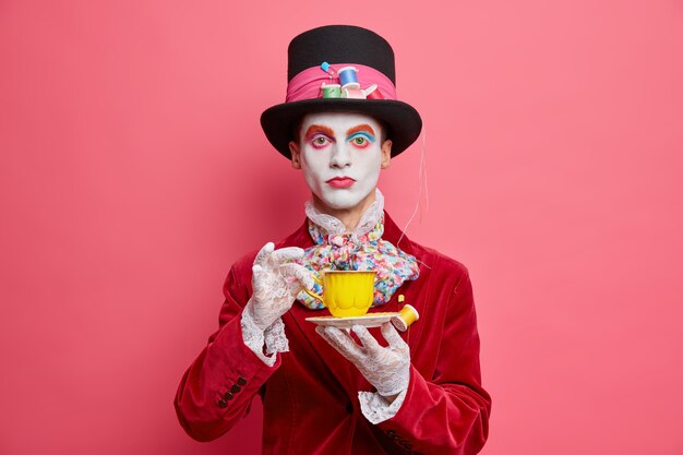 Serious fictional character hatter with colorful makeup drinks coffee has white face dressed in halloween costume looks seriously at camera isolated on pink wall