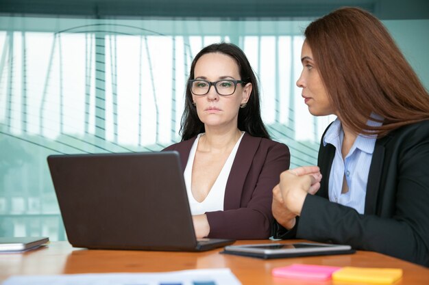 Serious focused business women discussing project and using laptop while sitting at meeting table.