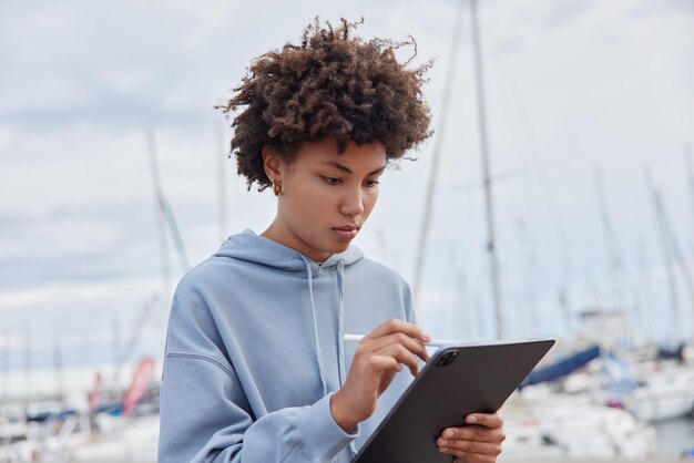 Serious focused creative curly woman makes drawings at digital tablet uses stylus pen wears casual sweatshirt poses againt blurred port background during daytime has inspiration draws sketch