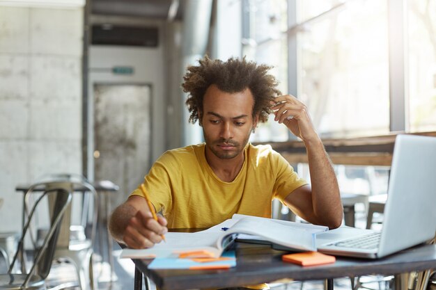 ラップトップコンピューターを使用して、黄色のtシャツを着た深刻な集中アフリカ大学生、家の割り当てを行う、忙しい練習帳に書き込む、早朝に空のコワーキングスペースに座っている