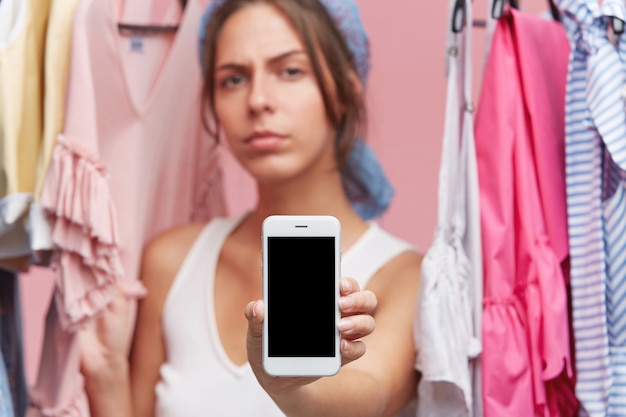 Free photo serious or frustrated young european female standing among multicolored clothes at closet or dressing room in clothing store, holding cell phone with copy space black screen for your information