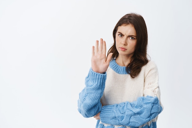 Serious girl frowning and showing block gesture, tell to stop or refuse from something bad, forbid an action, white wall