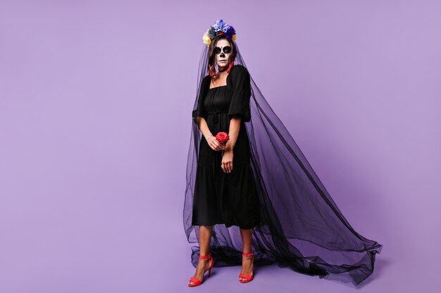 Serious girl posing as black widow on Halloween. Photo of model wearing crown of flowers and holding rose.