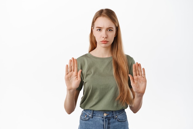 Foto gratuita ragazza seria che alza le mani per bloccare, dire di no, disapprovare l'azione e rifiutare qualcosa di brutto, accigliata dispiaciuta, rifiutando l'offerta, in piedi contro il muro bianco