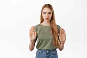 Free photo serious girl raising hands to block, say no, disapprove action and refusing something bad, frowning displeased, rejecting offer, standing against white wall