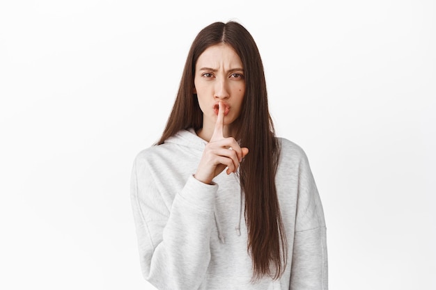 Serious girl tell to be quiet. Woman shushing and making taboo sign, keep secret, behave yourself, silence please, standing against white background
