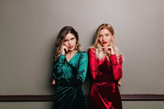 Serious girls in velvet clothes standing. Lovely slim ladies posing together on dark wall.