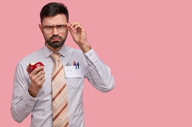 Серьезный мрачный мужчина-босс в строгой одежде держится за очки, кусает вкусное красное яблоко