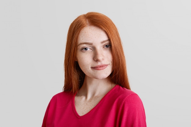 Serious good looking freckled young female model, has freckles on face, long straight red hair, dressed casually, looks with mysterious expression into camera, isolated over white wall.