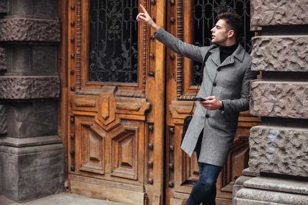Serious good-looking man standing near old building using cell phone, and catching taxi putting finger up