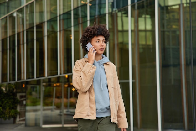 Serious good looking teenage girl makes telephone call concentrated into distance enjoys walking in city during pastime wears casual clothes poses against glass building. Urban lifestyle concept