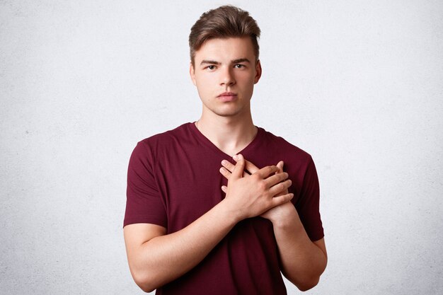 Serious good natured male keeps hands on heart