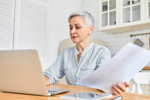 Serious gray haired mature businesswoman sitting at dining table using laptop for remote work, holding papers. Retired female making online payments via portable computer. Age, technology, occupation