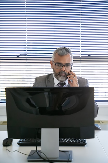 Foto gratuita grave uomo d'affari dai capelli grigi che fa chiamata sul telefono cellulare durante l'utilizzo del computer sul posto di lavoro in ufficio. vista frontale. comunicazione e concetto di multitasking